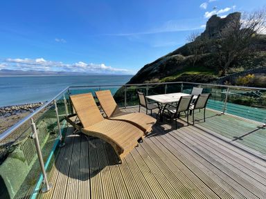 An outdoor deck with two lounge chairs and a dining table with four chairs overlooking the sea and a hillside at Awel Y Mor in Criccieth