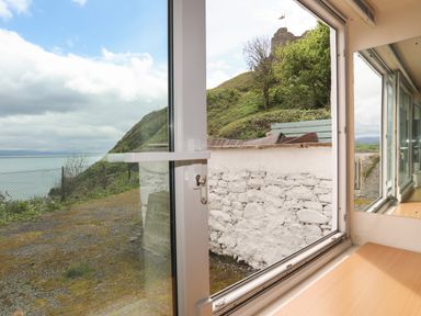 A view through a window showing a stone wall, a hill with trees, and water in the distance at Awel Y Mor in Criccieth