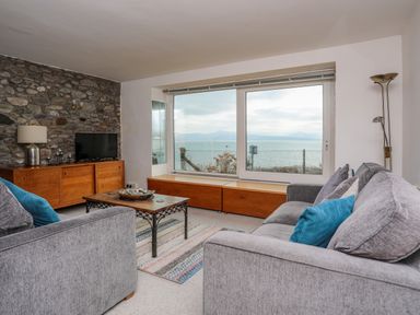 A living room with two gray sofas a wooden cabinet a flat screen tv and a large window with sea view at Awel Y Mor in Criccieth