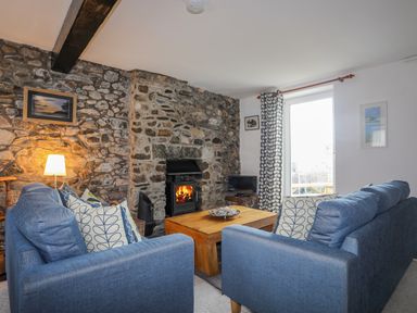 A living room with blue sofas a wooden coffee table a lit fireplace and stone walls at Awel Y Mor in Criccieth