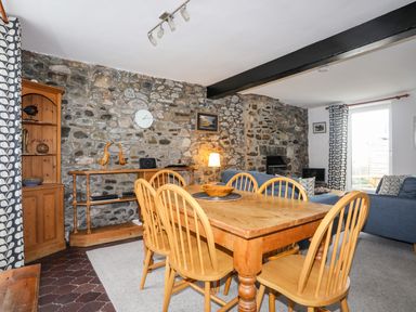 A dining area with a wooden table and chairs next to a seating area with blue sofas in a room with stone walls at Awel Y Mor in Criccieth