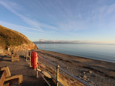A rocky beach with calm water and hills in the distance taken from a paved pathway with railings benches and a life buoy at Awel Y Mor in Criccieth