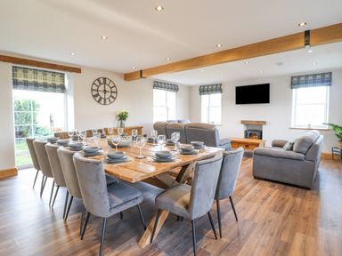 A dining area with a wooden table set with plates and glasses and a living area with gray sofas and a wall-mounted TV at Plas Iolyn in Pentrefoelas near Betws-Y-Coed