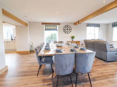 A dining area with a large wooden table set with plates and glasses next to a living room with gray sofas at Plas Iolyn in Pentrefoelas near Betws-Y-Coed