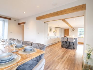 A dining area with a wooden table set with plates and glasses next to a kitchen with an island and bar stools at Plas Iolyn in Pentrefoelas near Betws-Y-Coed