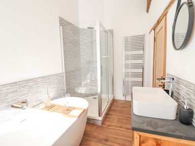 A bathroom with a bathtub with a wooden tray and glasses a glass shower enclosure a sink with a faucet and a round mirror a towel radiator and wooden flooring at Plas Iolyn in Pentrefoelas near Betws-Y-Coed