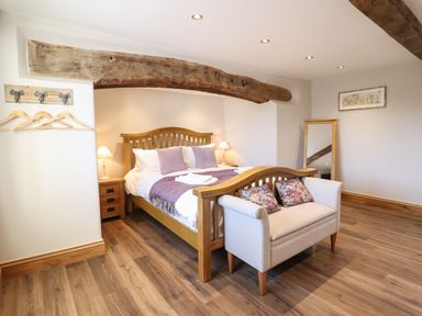 A bedroom with a wooden bed frame purple pillows and blankets a small sofa with floral cushions bedside tables lamps a large wooden beam on the ceiling and a full-length mirror at Plas Iolyn in Pentrefoelas near Betws-Y-Coed