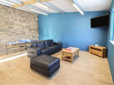A room with a black leather sofa and ottoman a wooden coffee table with a board game a stone wall a blue painted wall a wall-mounted TV and a ping pong table at Plas Iolyn in Pentrefoelas near Betws-Y-Coed
