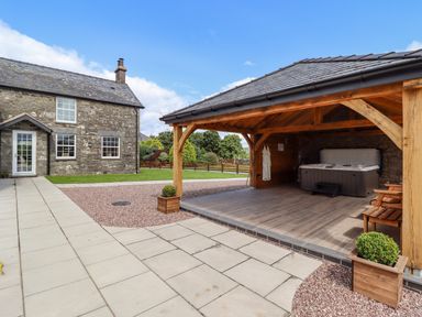 An outdoor area with a stone house, a wooden gazebo, a hot tub, a bench, potted plants, and a paved walkway at Plas Iolyn in Pentrefoelas near Betws-Y-Coed