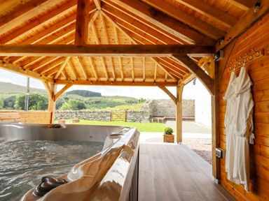 A covered outdoor area with a hot tub two white robes hanging on wooden wall and a stone wall garden at Plas Iolyn in Pentrefoelas near Betws-Y-Coed