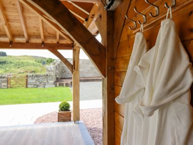 A covered wooden outdoor area with white robes hanging on hooks and a view of a garden with a stone wall and gate at Plas Iolyn in Pentrefoelas near Betws-Y-Coed
