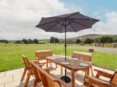 An outdoor wooden dining table with chairs and umbrella on a patio overlooking grassy fields and hills at Plas Iolyn in Pentrefoelas near Betws-Y-Coed