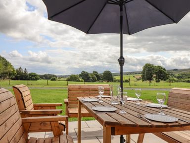 An outdoor wooden dining table set with plates and glasses under an umbrella with a green field and trees in the background at Plas Iolyn in Pentrefoelas near Betws-Y-Coed