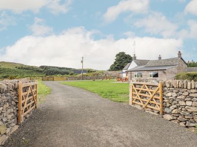 A gravel driveway with wooden gates and stone walls leading to a house with a lawn and outdoor seating at Plas Iolyn in Pentrefoelas near Betws-Y-Coed
