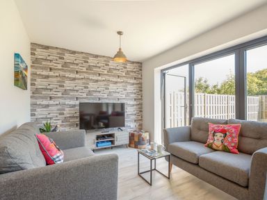 A living room with two grey sofas a television on a stand and glass doors opening to a fenced garden at Sea View in Llanfaelog