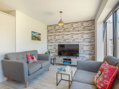 A living room with two grey sofas, a small table with cups, a TV on a stand against a patterned wall and a large window at Sea View in Llanfaelog