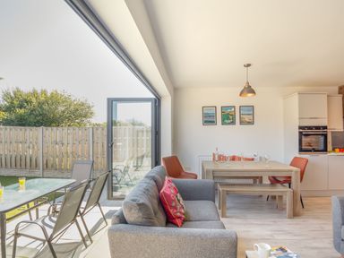 An open living and dining area with a gray sofa, wooden dining table, orange chairs, and glass doors leading to an outdoor patio at Sea View in Llanfaelog