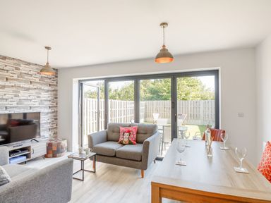 A living room with grey couches a wooden table and large glass doors opening to a garden at Sea View in Llanfaelog