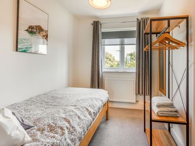 A bedroom with a single bed a wooden open wardrobe with hangers and folded towels a window with curtains and a wall picture at Sea View in Llanfaelog