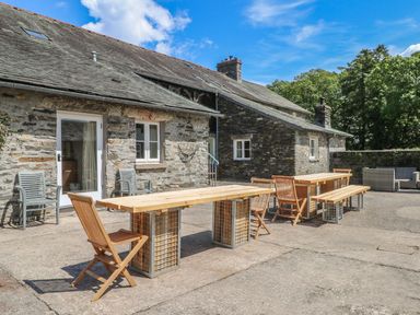 An outdoor patio area with wooden tables and chairs next to a stone building at Home Farmhouse Graythwaite near Hawkshead