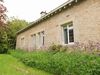 A stone exterior wall of a house with multiple windows and a flower bed with green plants and purple flowers in front at The Coach House in Shepherd's Dene near Corbridge