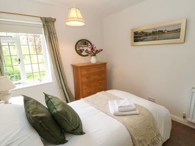 A bedroom with a bed with green pillows and folded towels a wooden chest of drawers with flowers and a round mirror by a window with curtains at The Coach House in Shepherd's Dene near Corbridge