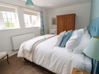 A bedroom with a bed with pillows and blankets a wooden wardrobe a chair a window with curtains and a radiator at The Coach House in Shepherds Dene near Corbridge