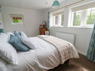 A bedroom with a bed, pillows, a wooden dresser with a mirror, a chair, and windows at The Coach House in Shepherd's Dene near Corbridge