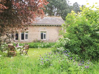 A garden with wooden chairs and a table surrounded by flowers and shrubs near a stone house