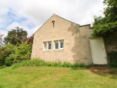 An exterior view of a stone building with three windows and a white door surrounded by grass and bushes at The Coach House in Shepherd's Dene near Corbridge