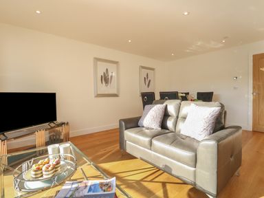 A living room with a leather sofa pillows a glass coffee table with mugs and pastries a flat screen tv and dining chairs at Harbourside Haven Apartment 2 in Weymouth