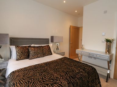 A bedroom with a bed featuring patterned pillows and blanket two bedside tables with lamps and a console table with flowers at Harbourside Haven Penthouse 1 in Weymouth