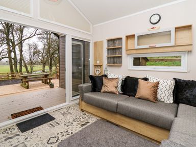 A living room with a sofa with cushions a wall clock and shelving and a sliding glass door opening to an outdoor picnic table at Woodland Escape in Rhayader