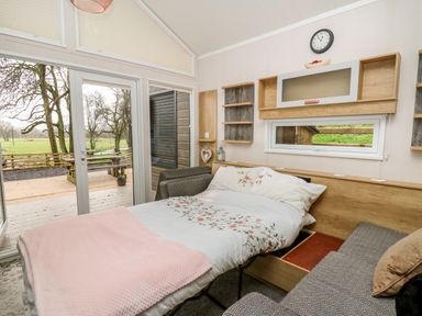 A bedroom with a bed, sofa, wall shelves, clock, and large glass doors opening to an outdoor area at Woodland Escape in Rhayader