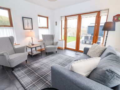 A living room with two chairs a sofa a side table a lamp and large glass doors opening to a garden at Cherry Blossom Cottage in Quilty County Clare