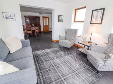 A living room with a gray sofa, two patterned armchairs, a side table with a lamp and clock, and a dining area with wooden furniture at Cherry Blossom Cottage in Quilty County Clare