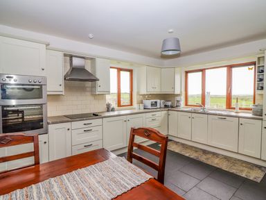 A kitchen with white cabinets a wooden table with chairs and large windows showing a landscape at Cherry Blossom Cottage in Quilty County Clare