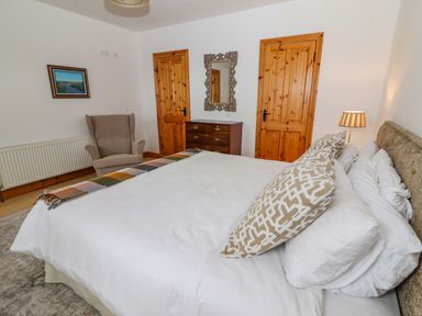 A bedroom with a bed white bedding patterned pillows a wooden dresser an armchair a mirror and two wooden doors at Cherry Blossom Cottage in Quilty County Clare