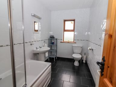 A bathroom with a bathtub shower unit sink toilet shelving unit and a window at Cherry Blossom Cottage in Quilty County Clare
