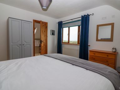 A bedroom with a bed a grey wardrobe a wooden chest of drawers a window with blue curtains and an open wooden door at Cherry Blossom Cottage in Quilty County Clare