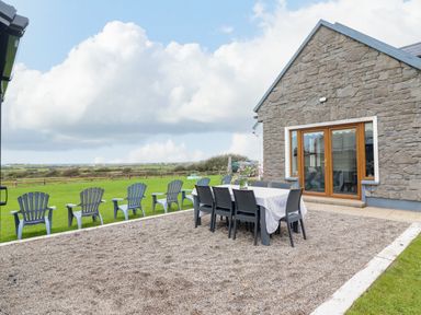 A stone house with wooden doors leading to a patio with a dining table and chairs and plastic chairs on grass at Cherry Blossom Cottage in Quilty County Clare