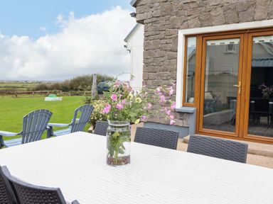 An outdoor patio with a table holding a vase of flowers and several chairs near a house with glass doors at Cherry Blossom Cottage in Quilty County Clare