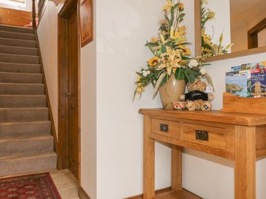 A hallway with a carpeted staircase wooden door a wooden table with a flower arrangement a teddy bear and brochures at Meadowside in Goonhavern