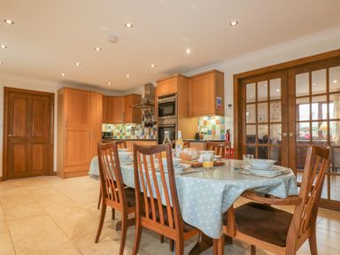 A kitchen with wooden cabinets a dining table with chairs and tableware and glass doors leading to another room at Meadowside in Goonhavern