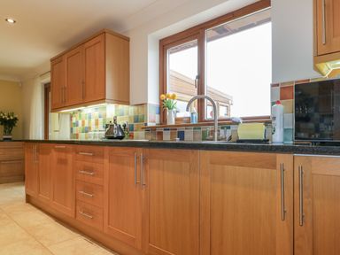 A kitchen with wooden cabinets and drawers a window above the sink with cleaning supplies and a kettle on the counter at Meadowside in Goonhavern