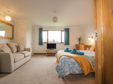 A bedroom with a bed covered in a patterned comforter and a mustard throw a beige sofa with two pillows a wooden dresser a window with teal curtains and a small TV on a wooden stand at Meadowside in Goonhavern