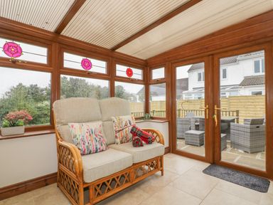 A sunroom with a two-seater wicker sofa with cushions and a blanket by wooden framed windows and glass doors at Meadowside in Goonhavern