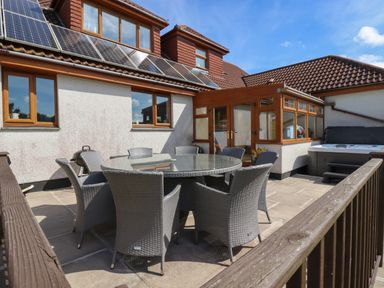 An outdoor patio with a round glass table and grey wicker chairs next to a house with solar panels and a hot tub at Meadowside in Goonhavern