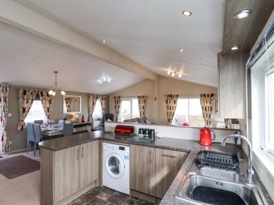 An open kitchen and dining area with wooden cabinets a washing machine a stainless steel sink red kettle and patterned curtains at Two Little Ducks in Cayton