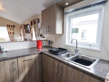 A kitchen with wooden cabinets a double sink a red kettle and windows with curtains at Two Little Ducks in Cayton